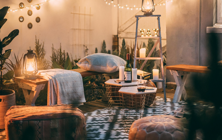 Cozy outdoor patio setup with warm string lights, lanterns, and a cozy candle on a wooden table—surrounded by cushions, blankets, and a bottle of wine, creating a relaxing evening atmosphere.
