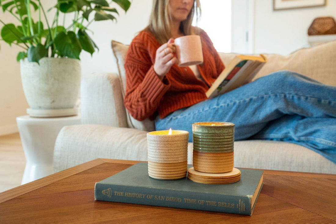Woman relaxing on a couch with a mug and book, with two ReCandle Co. refillable candles on the coffee table—showcasing the best candles for home in handmade ceramic vessels.