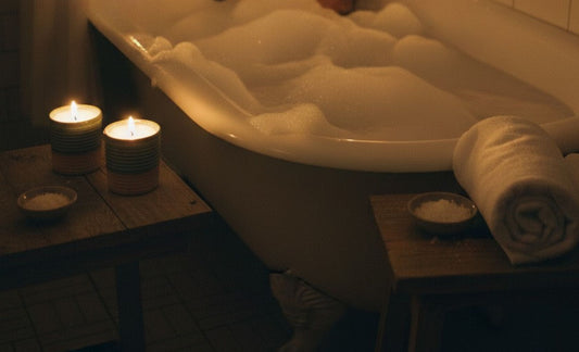 A cozy, nighttime lifestyle photo of a bubble bath in a dimly lit bathroom. Two ReCandle Co ceramic candles glow on a wooden side table in the foreground, while a person relaxes in a clawfoot tub in the soft, warm background.