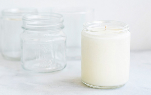 A white soy candle in a clear glass jar placed beside several empty glass jars on a light background, illustrating common single-use candle containers often reused or discarded