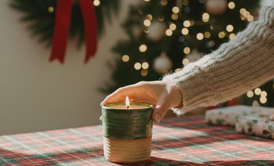 A person's hand reaching toward a lit, green and tan ceramic refillable candle on a red plaid tablecloth, with a blurred Christmas tree and wreath in the background.