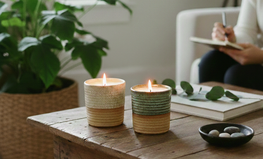 two lit ceramic candles on a wooden table in a sunlit room with a large plant and a person writing in the background.
