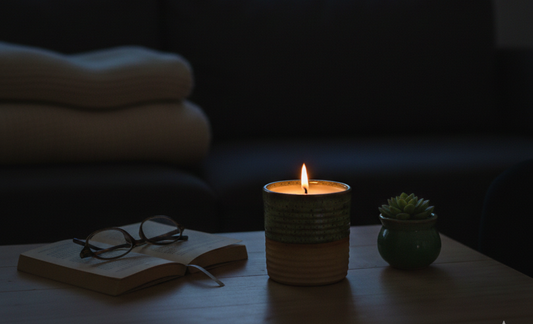 A cozy, nighttime lifestyle photo of a lit ceramic candle on a wooden table, alongside an open book, reading glasses, and a small succulent, with a soft-focus couch in the background.