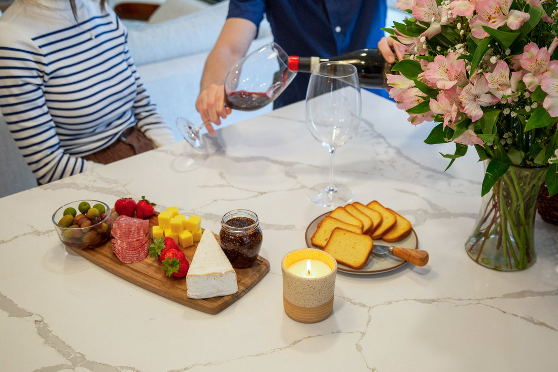 Candle lit on a table beside wine, cheese, and flowers during a cozy gathering.