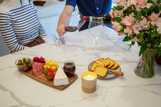 Candle lit on a table beside wine, cheese, and flowers during a cozy gathering.