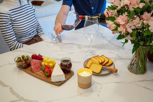 Candle lit on a table beside wine, cheese, and flowers during a cozy gathering.