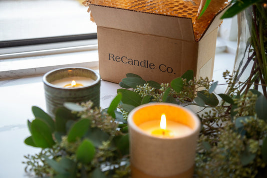 Two lit candles in ceramic vessels surrounded by greenery, with a ReCandle Co. shipping box in the background on a windowsill.