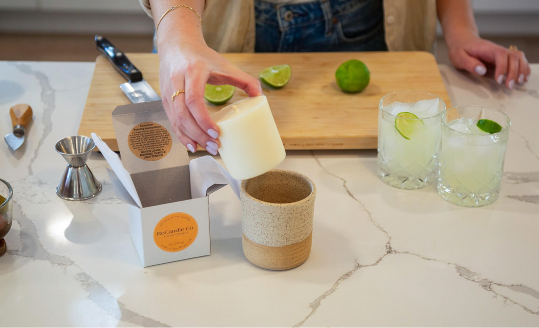 A person placing a ReCandle Co. candle refill into a ceramic vessel on a kitchen counter next to two lime cocktails.