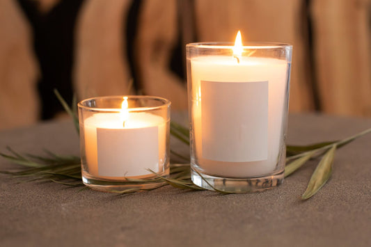 Two lit candles in clear glass jars—one small and one large—displayed side by side on a stone surface with green leaves, illustrating different candle jar sizes.