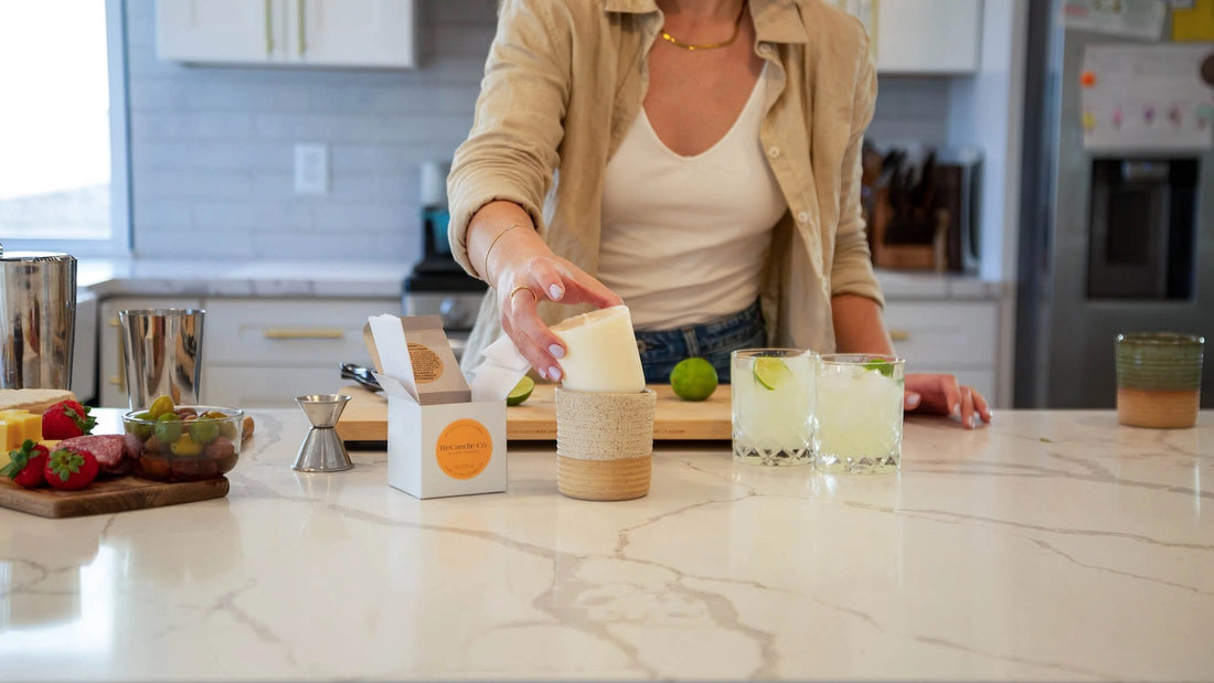 A person placing a soy wax refill into a reusable candle jar on a kitchen countertop, demonstrating how to reduce waste by reusing candle containers