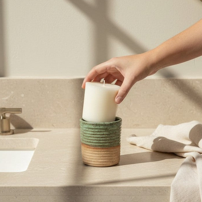 A hand places a white pillar candle into a stacked set of ceramic holders, including a green ribbed top and a natural wood base, set on a beige bathroom vanity countertop.