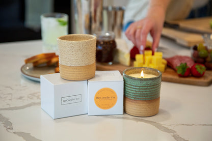 Two ReCandle Co. refillable candles sit on and next two soy wax candle refill boxes on a kitchen counter top, and a someone grabs a piece of cheese off a charcuterie board behind the candles.