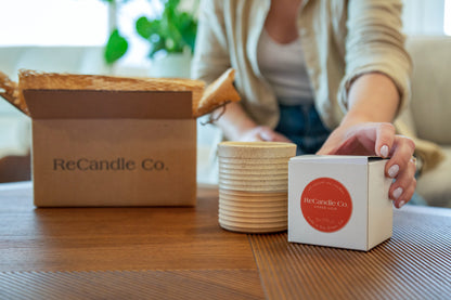 Someone grabs a ReCandle Co. soy wax refill box, next to the ReCandle Co. refillable candle vessel. The refillable candle is on a coffe table, next to an empty soy wax refillable candle box and a 100% recyclable ReCandle Co. packaging box.