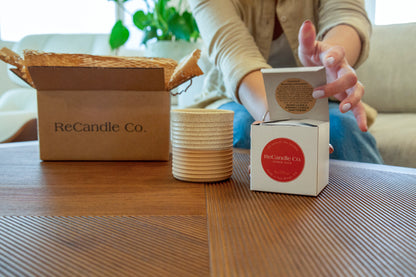 Someone opens a ReCandle Co. soy wax refill box next to an empty refillable candle vessel. The refillable candle is on a coffee table, next to an empty soy wax refillable candle box and a 100% recyclable ReCandle Co. packaging box.