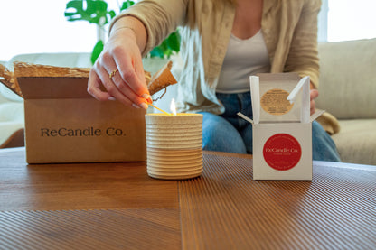 Someone lights a ReCandle Co. refillable candle that's placed on a coffee table, next to an empty soy wax refillable candle box and a 100% recyclable ReCandle Co. packaging box.
